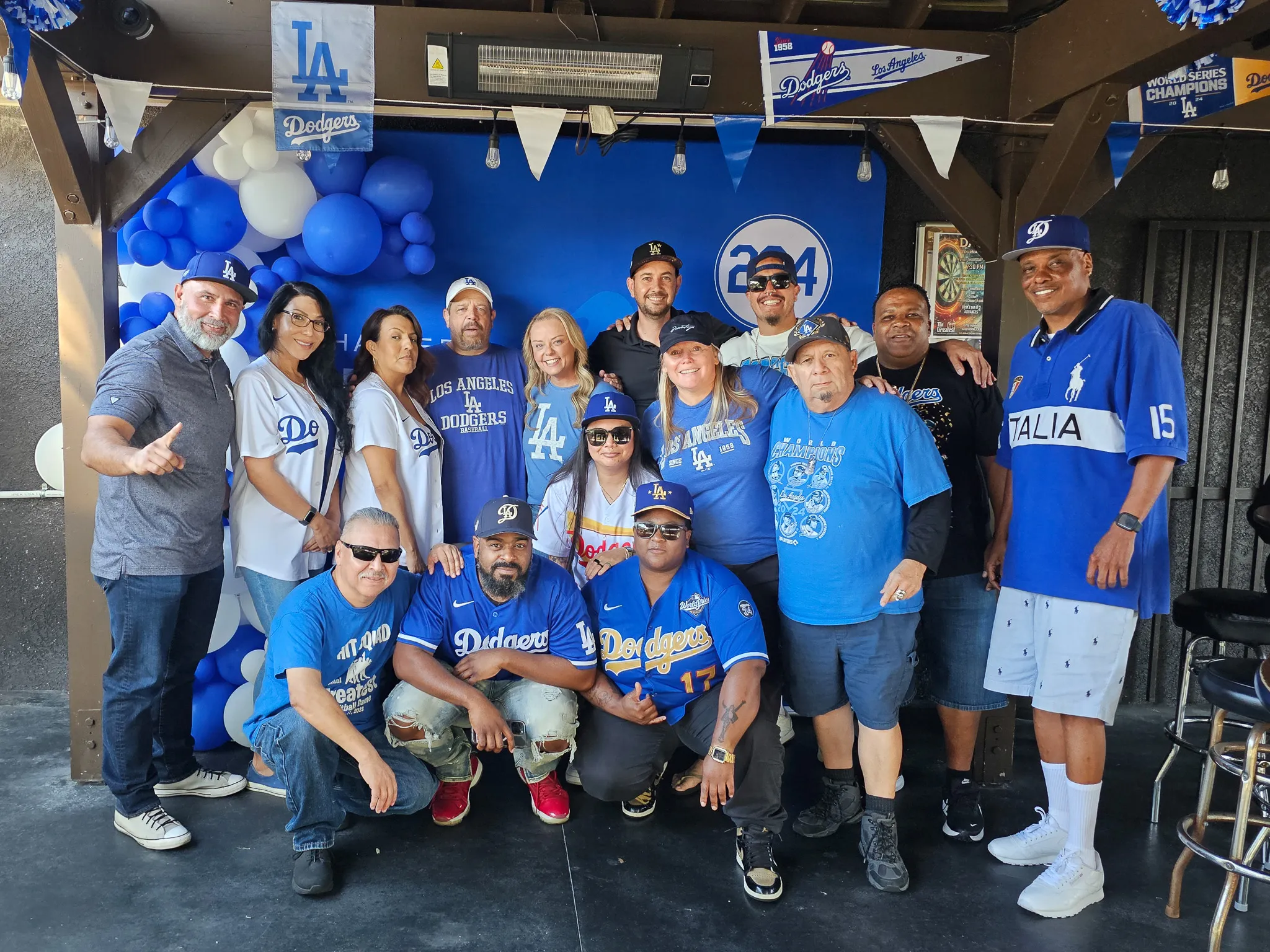 Group photo of Dodgers fans at The Greatest Sports Bar in Hawthorne in front of the Chapter 294 blue backdrop and blue-and-white balloon arch. Fans in standing and crouching rows wear Dodgers jerseys and gear. LA and Dodgers pennants and a World Series Champions banner are displayed on the walls.